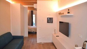 Ett kompakt vardagsrum med en soffa, en platt-TV som är monterad på väggen, en liten hylla med en flaska och en dörr som leder till ett annat rum.