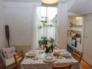 A dining room with a table set for four, a window with a view of the outside, and a kitchen visible through an open door. - Shipwrights Cottage (Teignmouth)