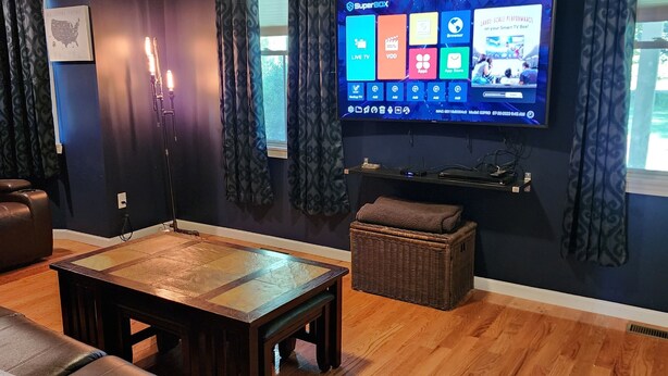 Een woonkamer met een flatscreen televisie aan de muur, een houten salontafel, een bruine bank en een rieten poef.