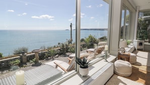 Habitació amb finestra gran que ofereix vista al mar, amb sofà, taula petita i una planta en test.