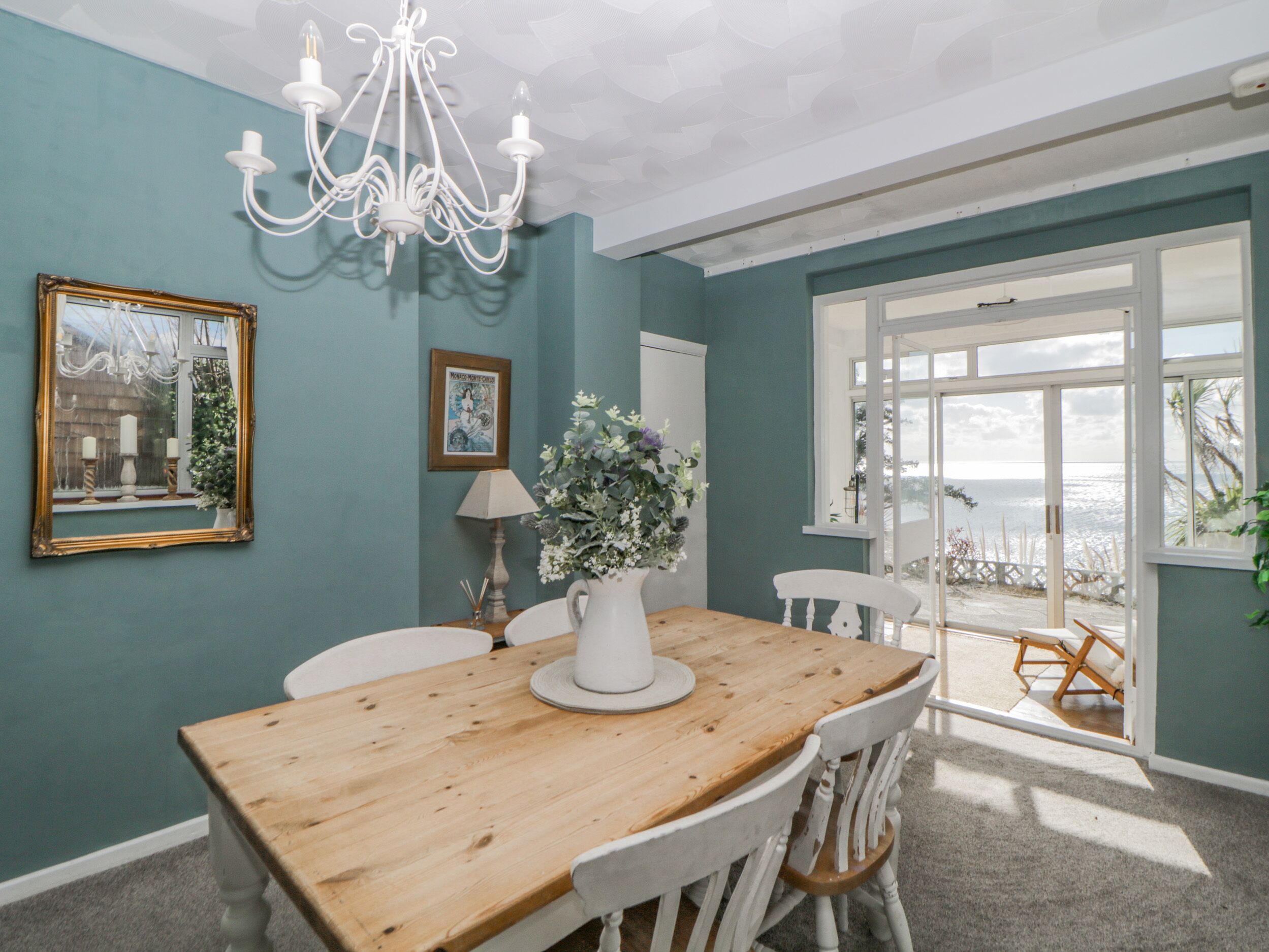 A dining area with a wooden table, white chairs, a chandelier, and a view of the sea through large windows.