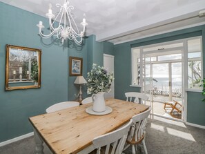 Un comedor con mesa de madera, sillas blancas, una araña y vista al mar a través de amplios ventanales.
