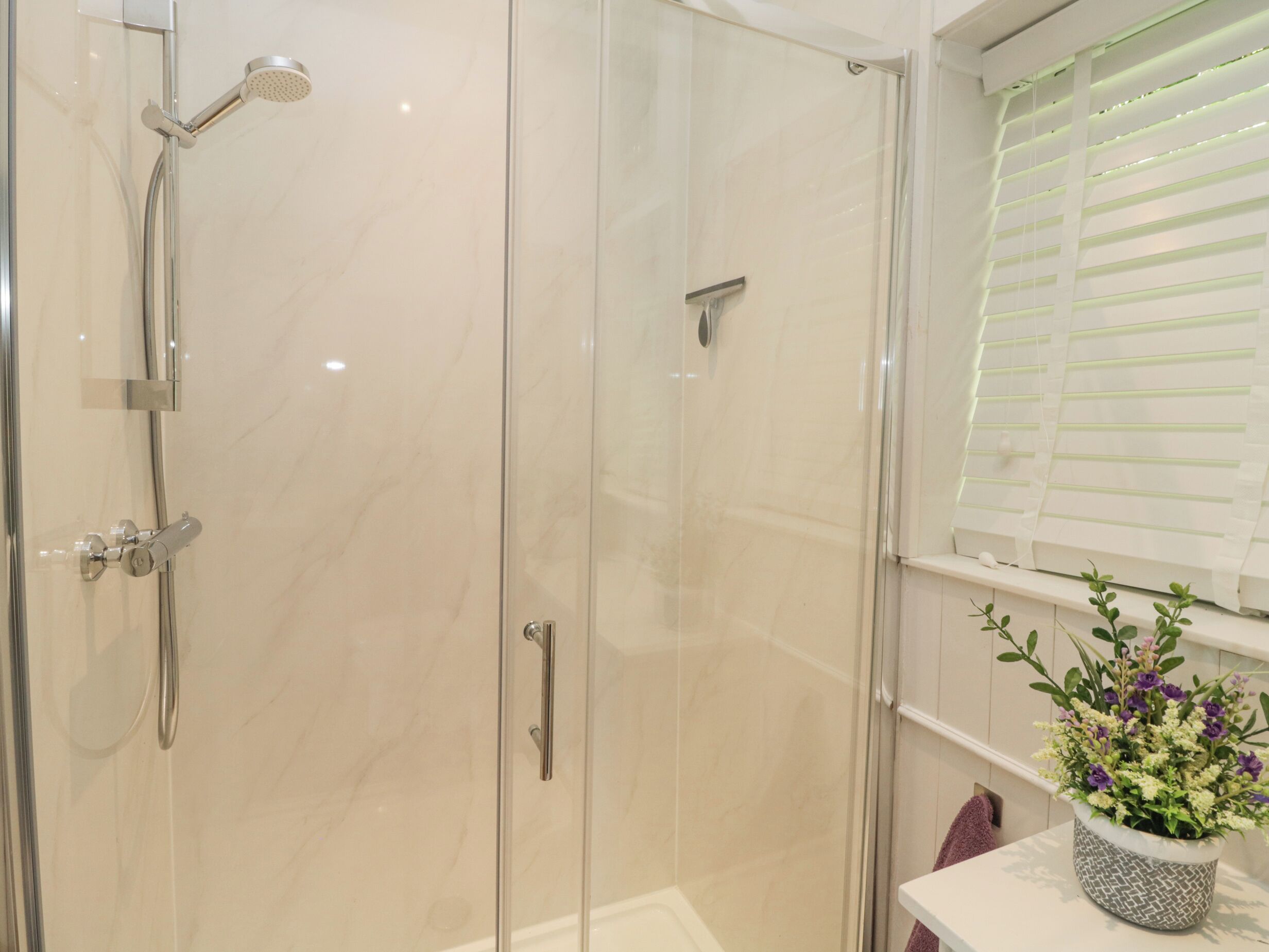 Un baño moderno con ducha envuelta en vidrio, tocador con lavamanos y ventana con persianas.