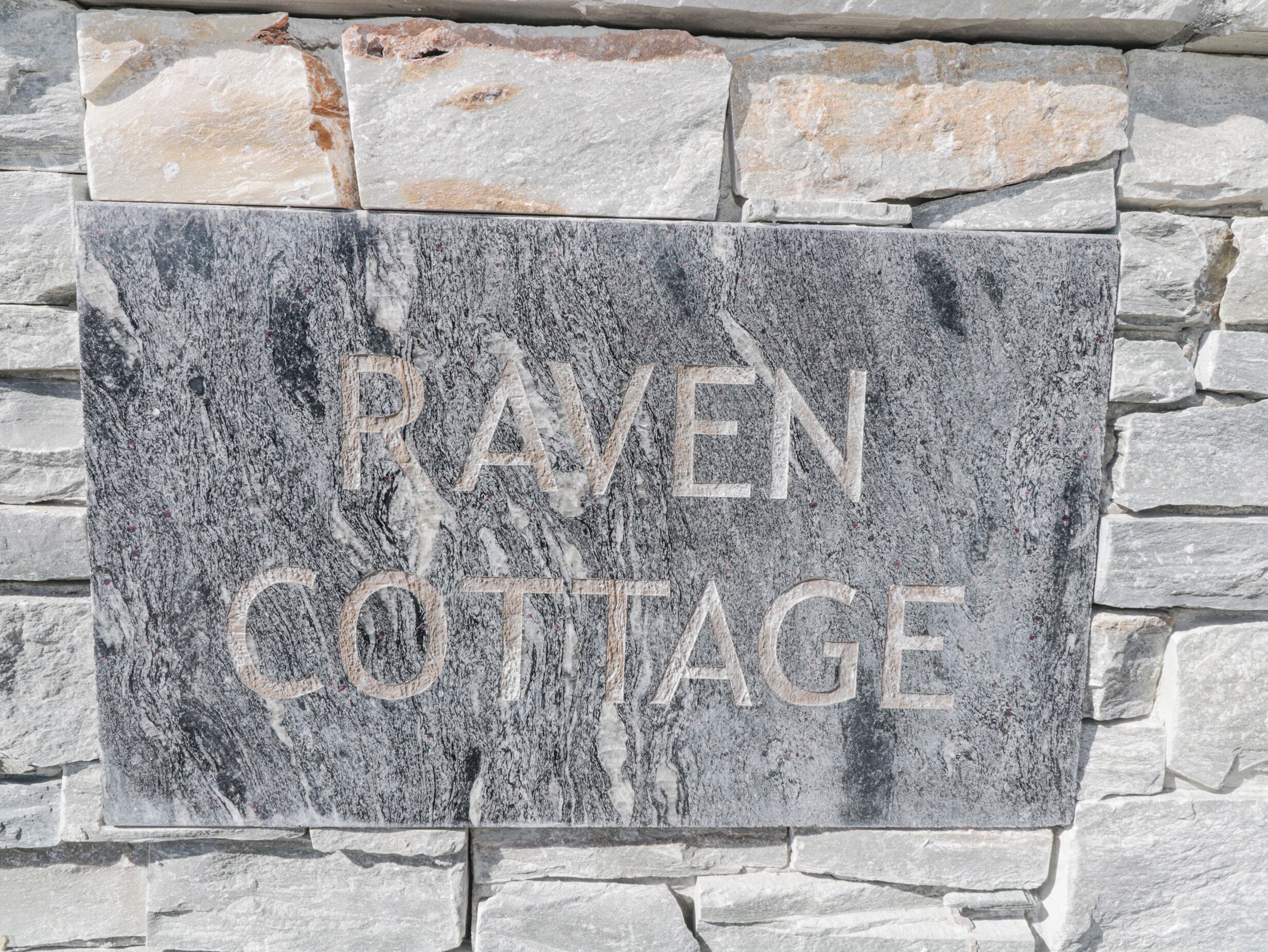 A stone sign with the text "RAVEN COTTAGE" against a stone wall background.