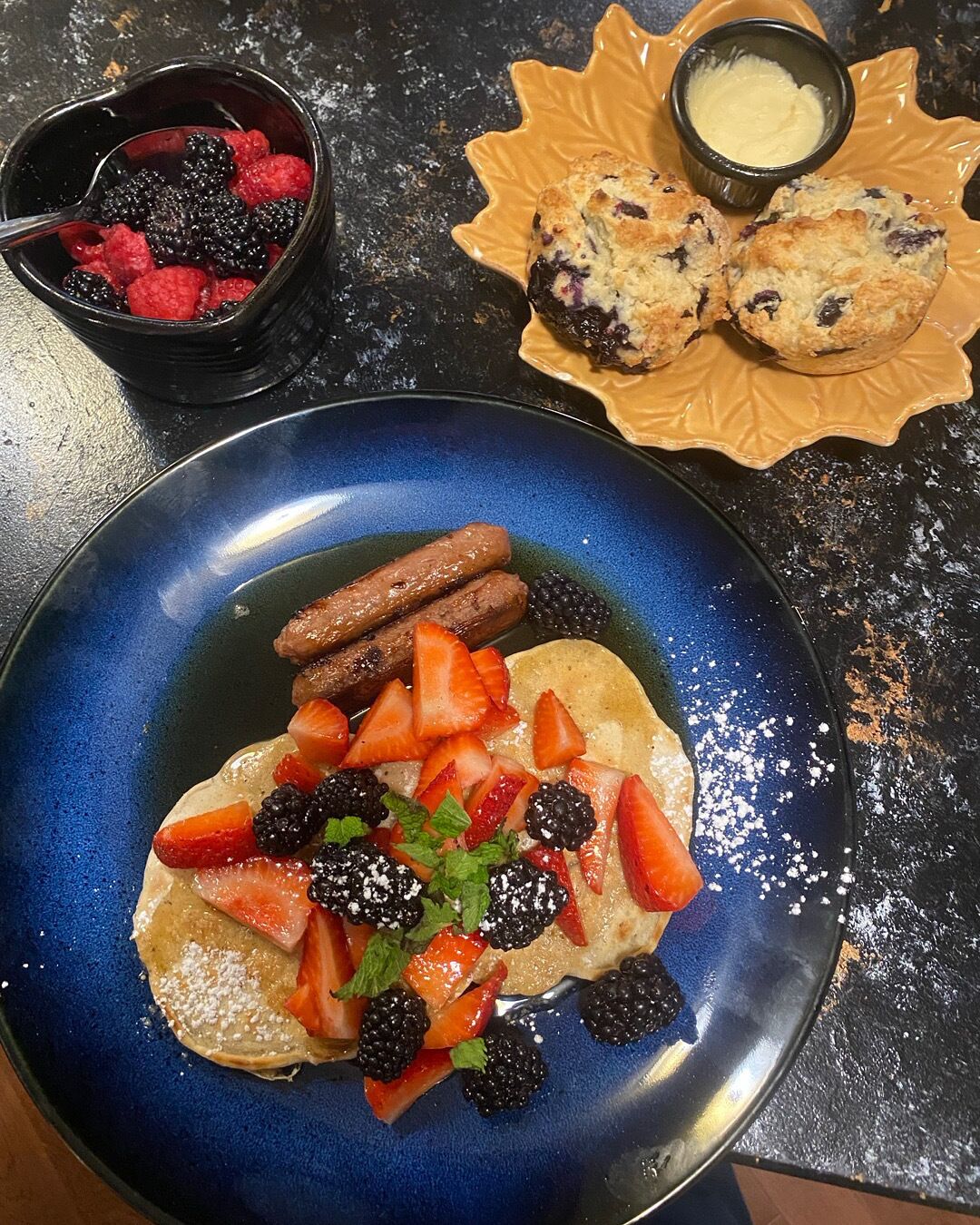 A plate of pancakes topped with strawberries, blackberries, and mint, accompanied by a bowl of berries and a plate of scones.