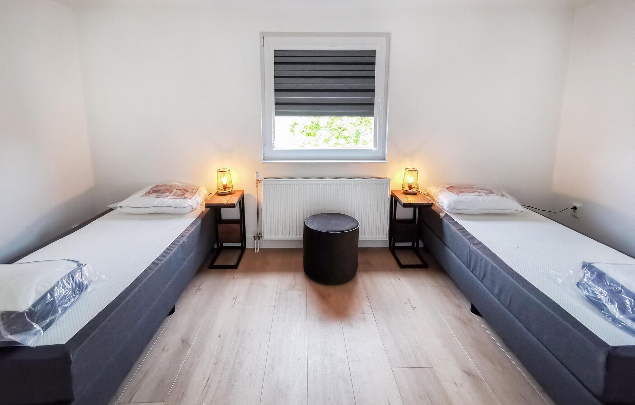 A room with two single beds, a small round stool, a radiator, and a window with blinds.