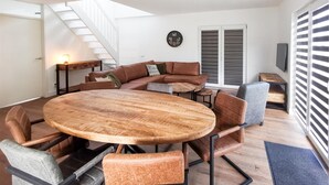 Una sala moderna con una mesa de comedor de madera rodeada de sillas, un sofá y una mesa de centro. Se ve una escalera al fondo, que sube a un nivel superior.