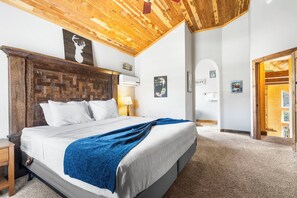 Ein Schlafzimmer mit Holzbalkendecke, einem Bett mit weißen Leinen und einer blauen Decke, einem Nachttisch und einer Wand-Klimaanlage.