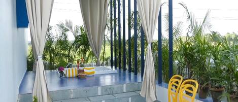 A covered outdoor area with steps leading to a seating area with yellow chairs, potted plants, and a view of greenery.