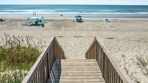 Une promenade en bois menant à une plage de sable où se trouvent une tente et un parasol bleu.