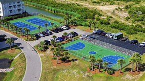 An aerial view of a sports complex with multiple tennis courts, parking areas, and a building adjacent to a body of water. - A Wave from it All by Brunswick Vacation Rentals: Beach Club w/ Pool, Elevator (Ocean Isle Beach)