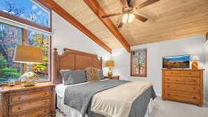 A bedroom with a wooden ceiling, a large bed with grey bedding, a wooden nightstand with a lamp, and a dresser. - Mountain Home near Skiing, Hiking, & More (Beech Mountain)