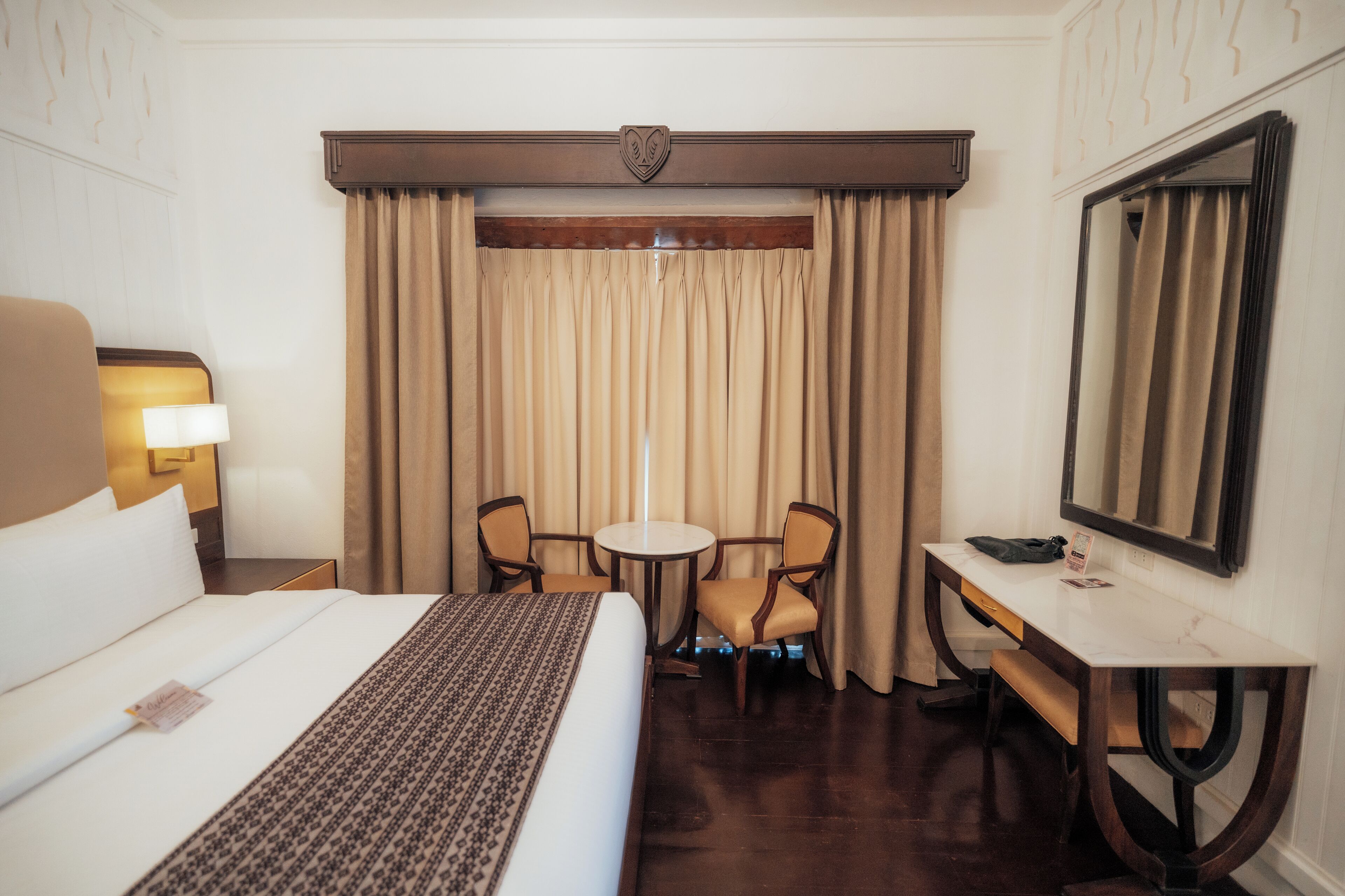 A hotel room with a large bed, a desk, two chairs, a round table, a mirror, and a window with curtains.