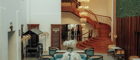 A spacious lobby with a geometric patterned floor, teal seating, a central table, and a staircase leading to an upper level.
