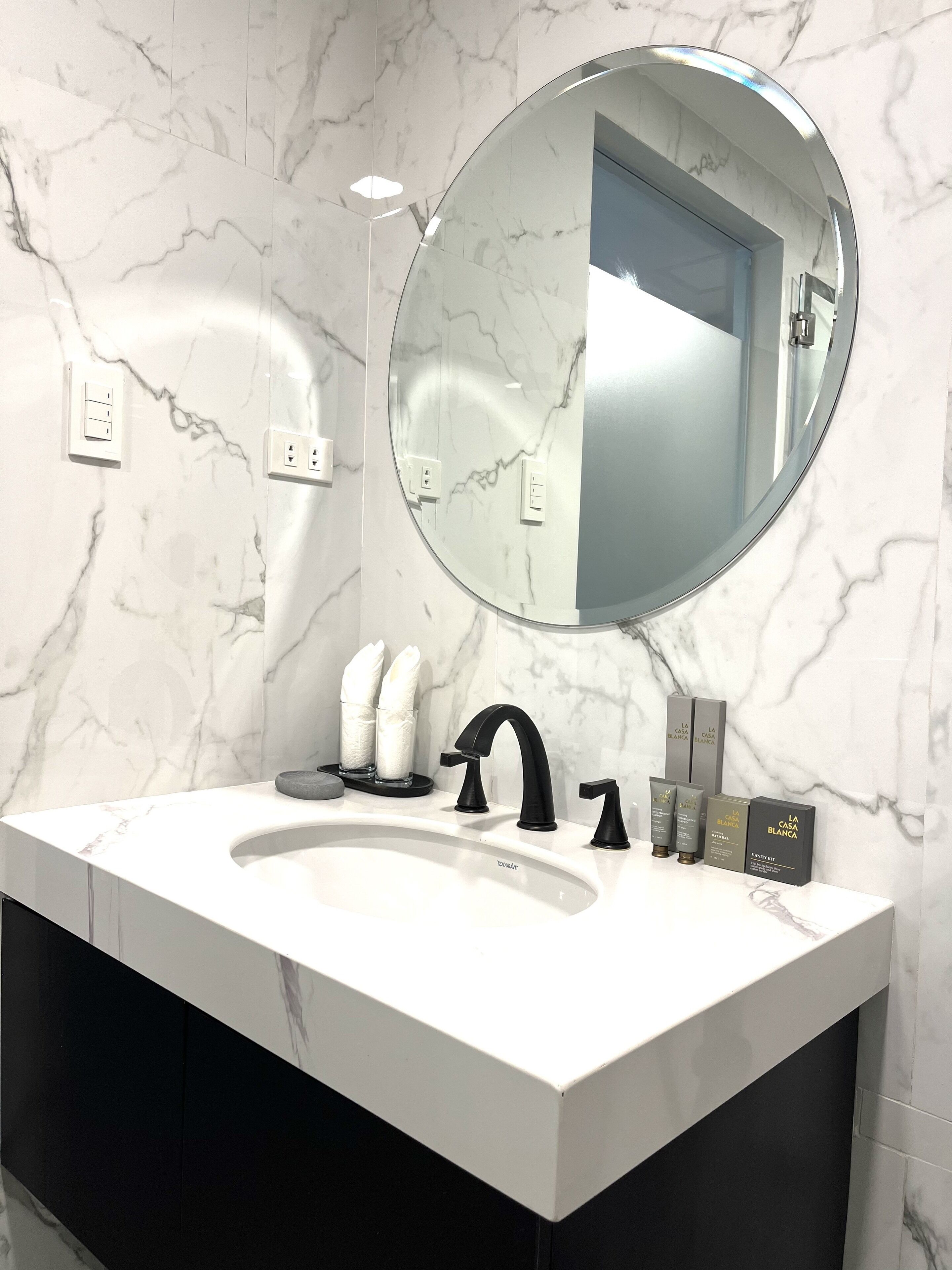 A modern bathroom with a marble sink, a round mirror, and a black faucet. There are two towel dispensers and a set of toiletries on the counter.