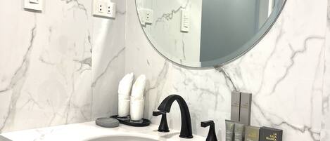A modern bathroom with a marble sink, a round mirror, and a black faucet. There are two towel dispensers and a set of toiletries on the counter.