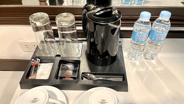 Ein Frühstücksbrett im Hotelzimmer mit zwei weißen Tassen, einer Kaffeemaschine, einem Wasserkocher, einer Wasserflasche und einem Zuckerbeutel.