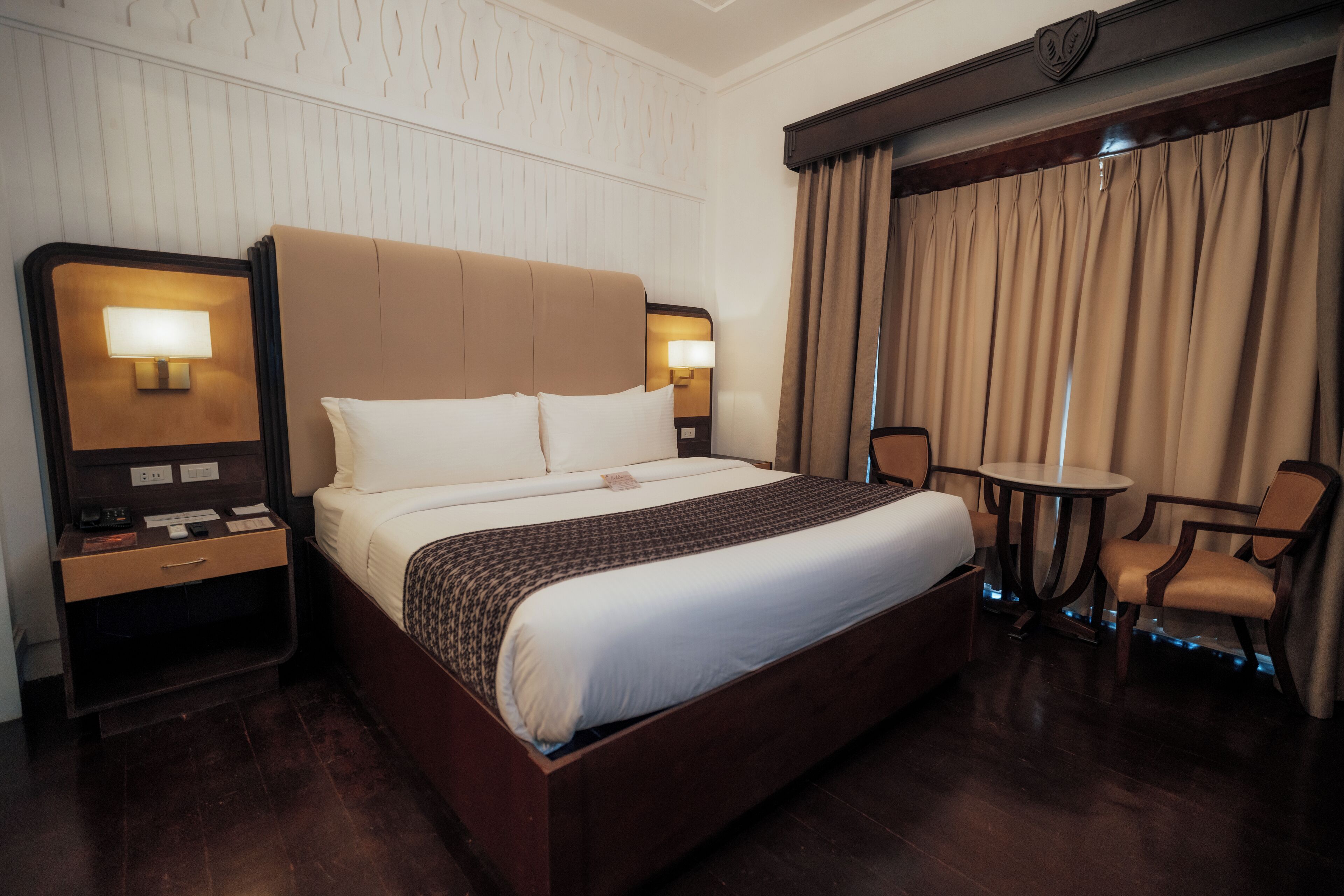 A hotel room with a large bed, two bedside tables with lamps, a small round table, and a chair.