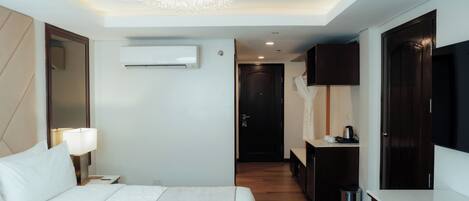 A hotel room with a large bed, a bedside lamp, a chandelier, an air conditioning unit, a TV, and a small kitchenette.
