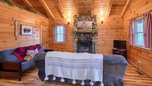 Ein gemütliches Blockhaus-Interieur mit Kamin aus Stein, Holzwänden, einer Couch und einem Fernseher.