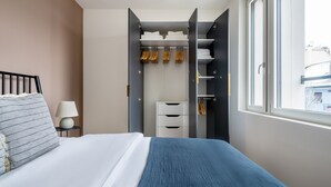Une chambre à coucher avec un lit, une table de chevet avec une lampe, une armoire contenant des vêtements et une fenêtre donnant sur des immeubles.