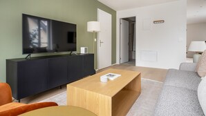 Un salon moderne avec un téléviseur à écran plat, un meuble sombre, une table basse en bois, un canapé gris et une porte donnant accès à une salle de bain.
