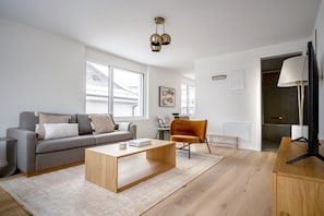 Ein modernes Wohnzimmer mit einem grauen Sofa, einem Holztisch und einem roten Sessel. Ein großes Fenster sorgt für natürliches Licht und einen Ausblick ins Freie.