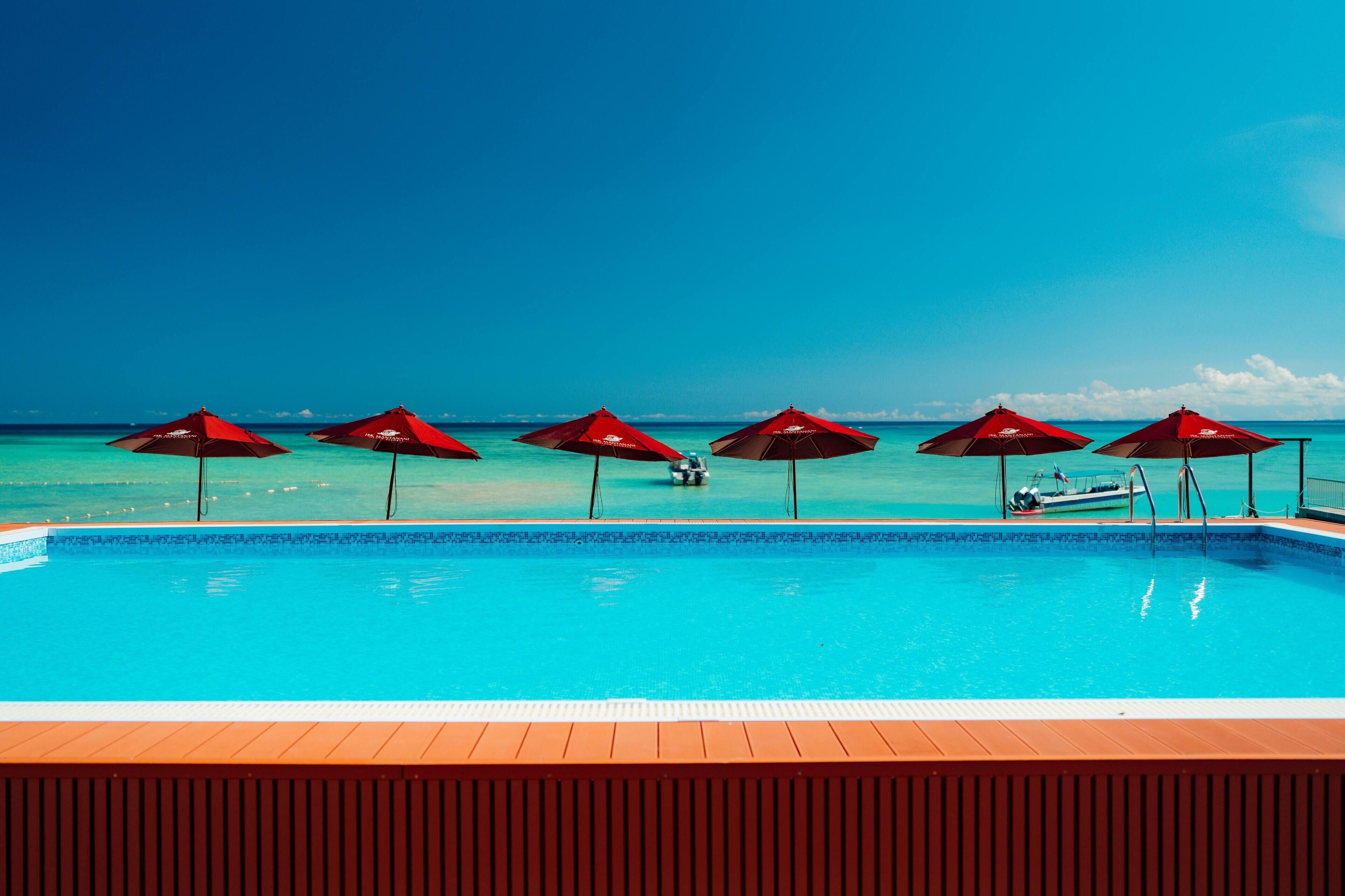 A swimming pool with six red umbrellas, clear blue water, and a boat in the background.