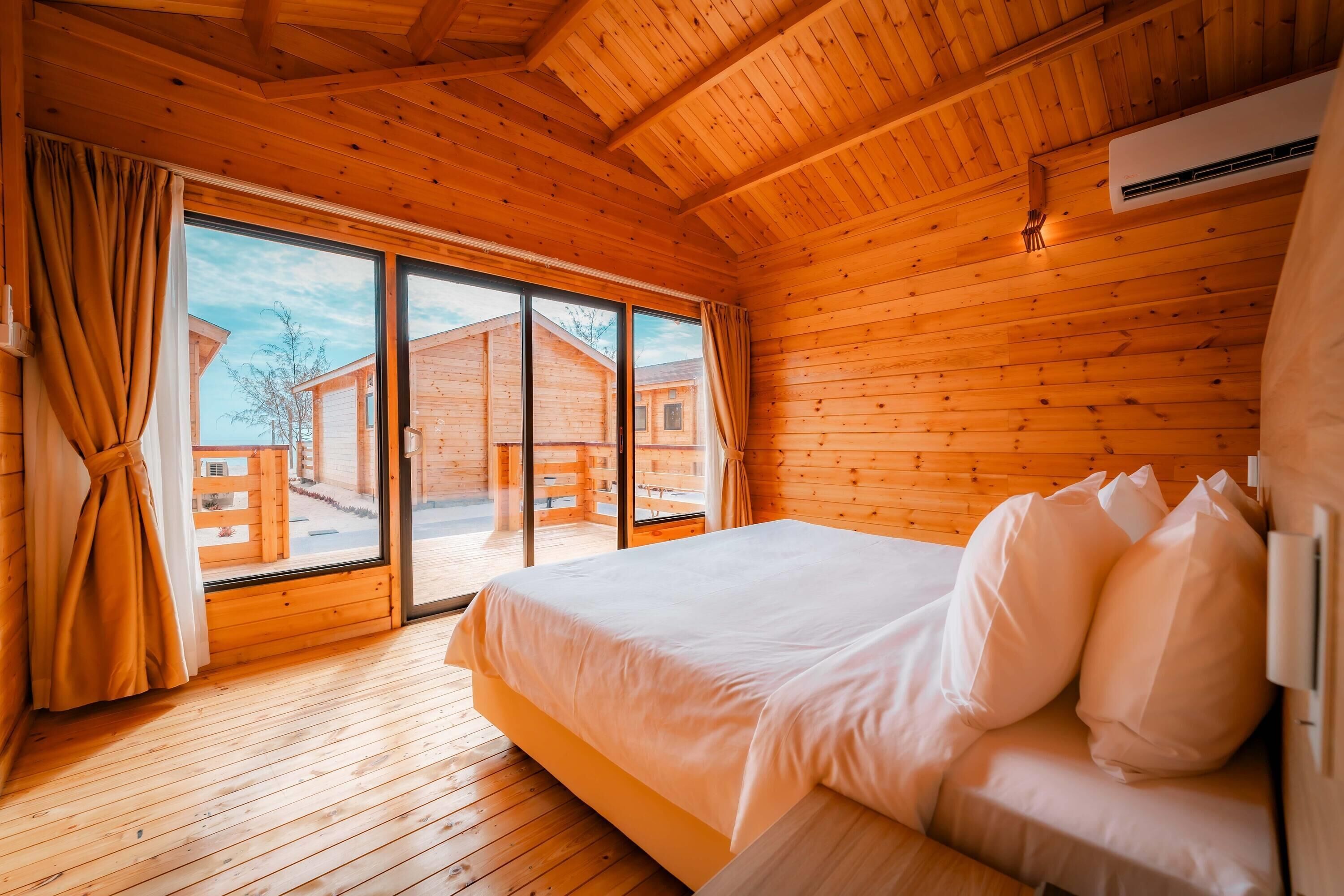A wooden cabin room with a bed, curtains, and a sliding glass door leading to an outdoor area.