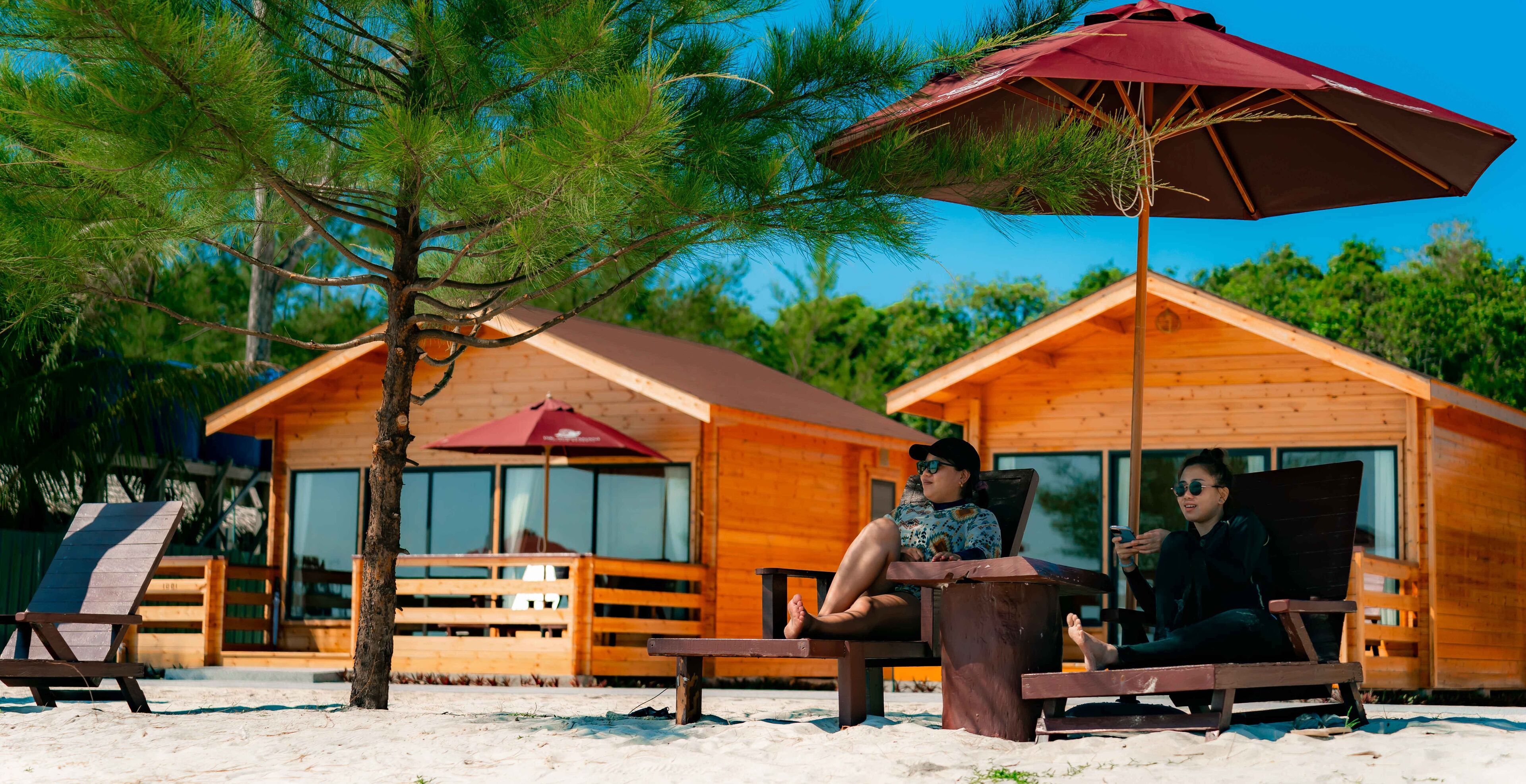 Uma cabana de madeira com vista para a praia, um guarda-sol vermelho e uma cadeira de lounge.