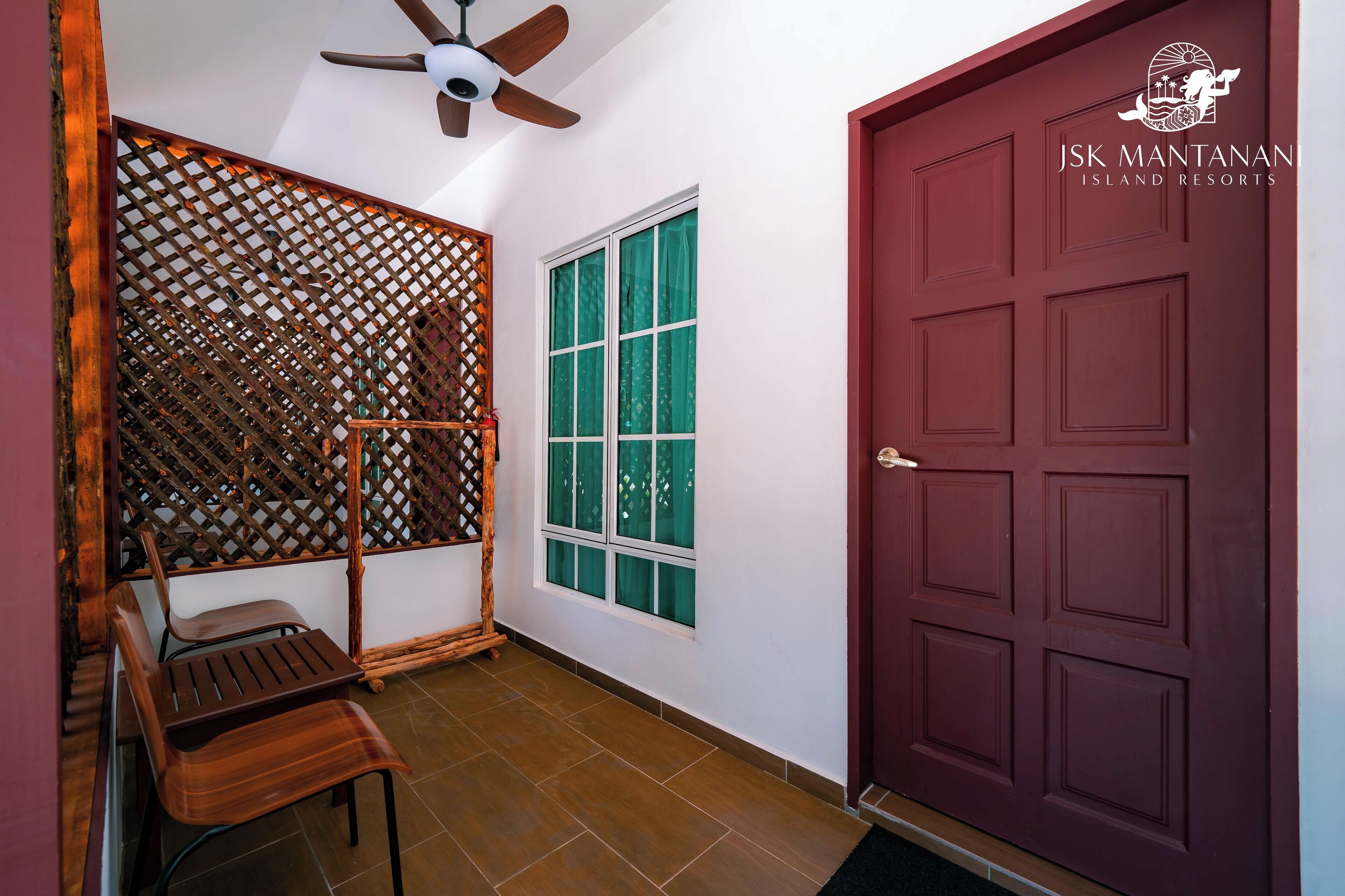 A room with a wooden chair, a table, a ceiling fan, a window with green curtains, and a red door.