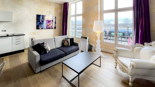 A living room with a sofa, a coffee table, a white armchair, a lamp, and a large window with curtains.