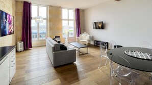 A modern living room with a sofa, armchair, and a dining table with chairs. There is a TV mounted on the wall and a large window with curtains.