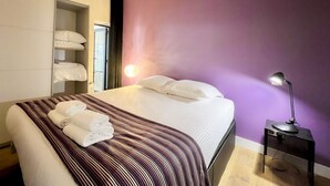 A neatly made bed with white linens and a striped blanket, a bedside lamp, and a shelf with folded towels.