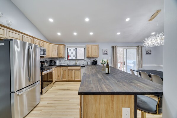A modern kitchen with wooden cabinets, a stainless steel refrigerator, a marble countertop, and a dining area with chairs.