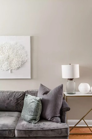 A modern living room with a grey sofa, a white abstract wall art, a white table lamp, and a small white vase.