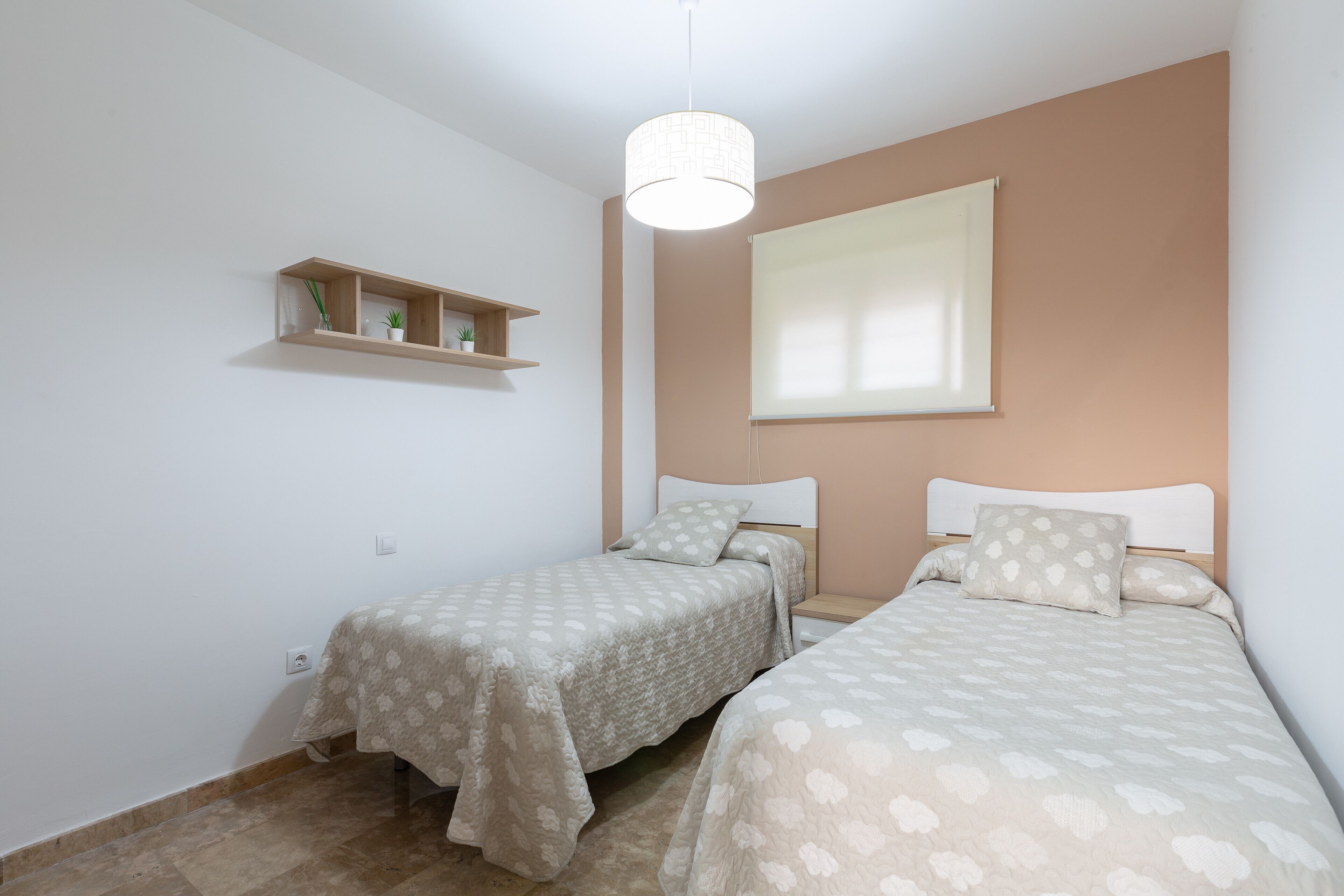 Een slaapkamer met twee bedden, een raam met zonwering, een plank met planten en een hangende lamp.