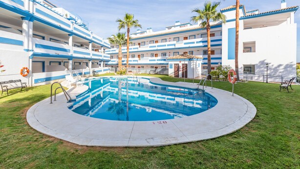 A hotel with a central swimming pool surrounded by palm trees and a well-maintained lawn. - CASA CARMEN - Wonderful apartment with shared pool and free WiFi. (Estepona)