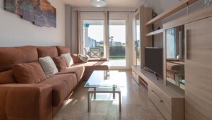 A modern living room with a brown sofa, a glass coffee table, a flat-screen TV, wooden shelves, and a large window with a view of the outdoors. - CASA CARMEN - Wonderful apartment with shared pool and free WiFi. (Estepona)