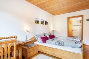 Una cama de madera con colchón blanco y ropa de cama floral, un escritorio de madera con silla, una mesita de noche y una lámpara fijada a la pared.