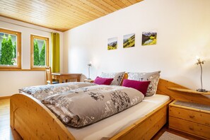 Een houten bed met een bloemenbedekking, een nachtkastje met een lamp, en uitzicht op bomen door twee ramen.