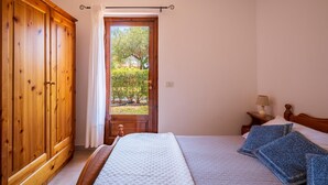 A bedroom with a wooden wardrobe, a bed with a quilt, a nightstand with a lamp, and a view of a garden through an open door. - 2 bedroom beautiful apartment in San Teodoro (San Teodoro)