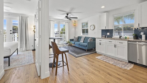 A spacious living room with a sofa, coffee table, and chairs, a kitchen with white cabinets and a sink, and a balcony with a view.