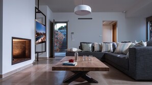 A modern living room with a fireplace, a large sofa, a coffee table, and a view of a cityscape through a large window. - Luxurious Sea View Villa in Agios Nikolaos (Agios Nikolaos)