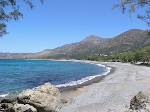 A beach with pebbles, a clear blue sea, and mountains in the background.