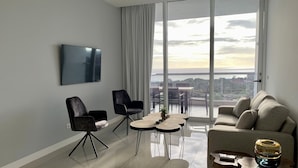 Un salon moderne doté d’une grande fenêtre, d’un téléviseur à écran plat, de deux fauteuils, d’un canapé et d’une table basse.
