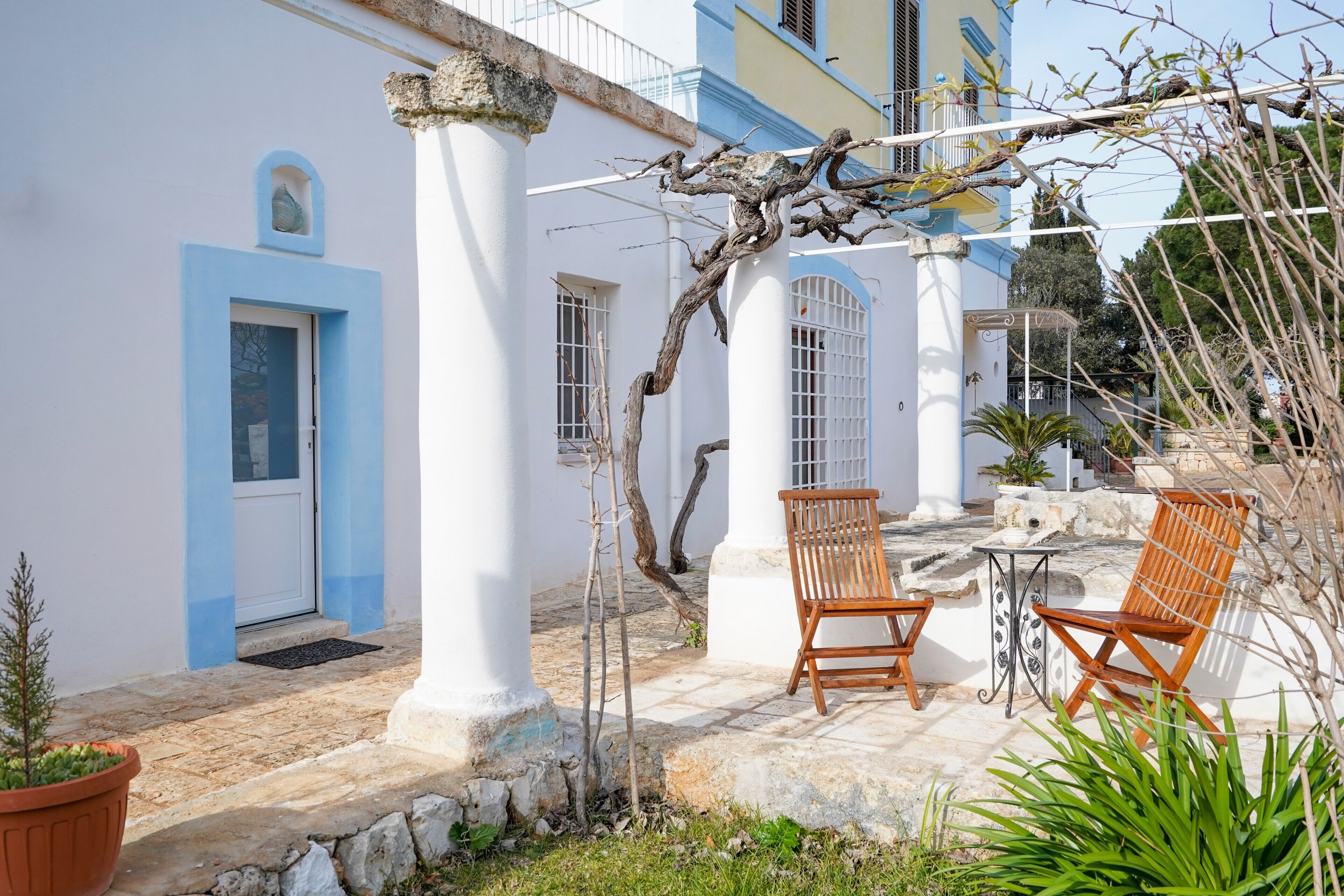 A courtyard with a white building, blue door, and columns, featuring two wooden chairs and a table.