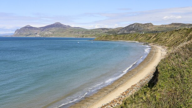 A coastal landscape with a sandy beach, rocky cliffs, and a clear blue sea. - Tir Eironwy (Pwllheli)