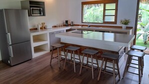 Une cuisine moderne dotée d’un îlot central, de tabourets de bar, d’un réfrigérateur, d’un four à micro-ondes et d’une fenêtre donnant sur un espace verdoyant.