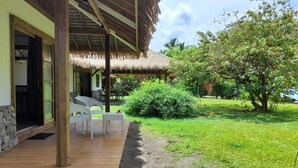 Une terrasse en bois avec des chaises blanches, une structure dotée d’un toit de chaume et un jardin luxuriant avec un grand arbre et des arbustes.
